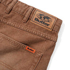 Close-up of the Distressed Brown Relaxed Denim Jeans by Butter Goods, showing a back pocket with a small orange tag and a large leather Butter logo patch above the waistband. Made from 100% cotton denim.