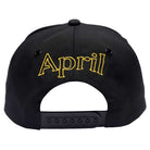 A black adjustable snapback hat from April Skateboards with "April" logo stylishly embroidered in gold above the strap.