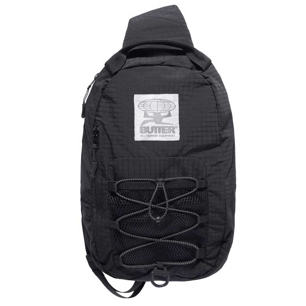 The Express Shoulder Bag Black Butter Goods features a sleek black design, top handle, front zipper pocket, crisscrossed elastic cord, and a gray patch with globe and "Butter Goods All Terrain Equipment" text—ideal for stylish versatility.