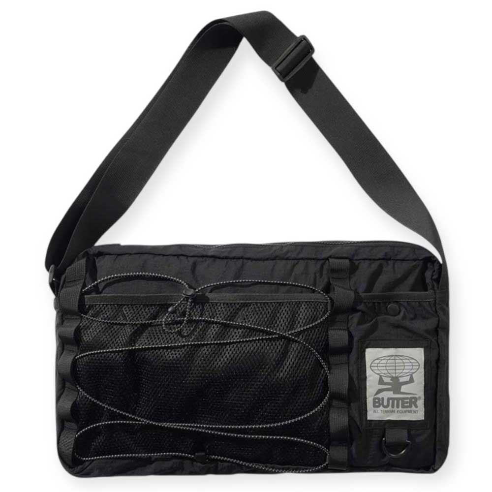 The Express Side Bag Black by Butter Goods features a wide adjustable strap, mesh front pocket, cord details, and a globe logo patch with "BUTTER" above smaller text. Made from nylon.