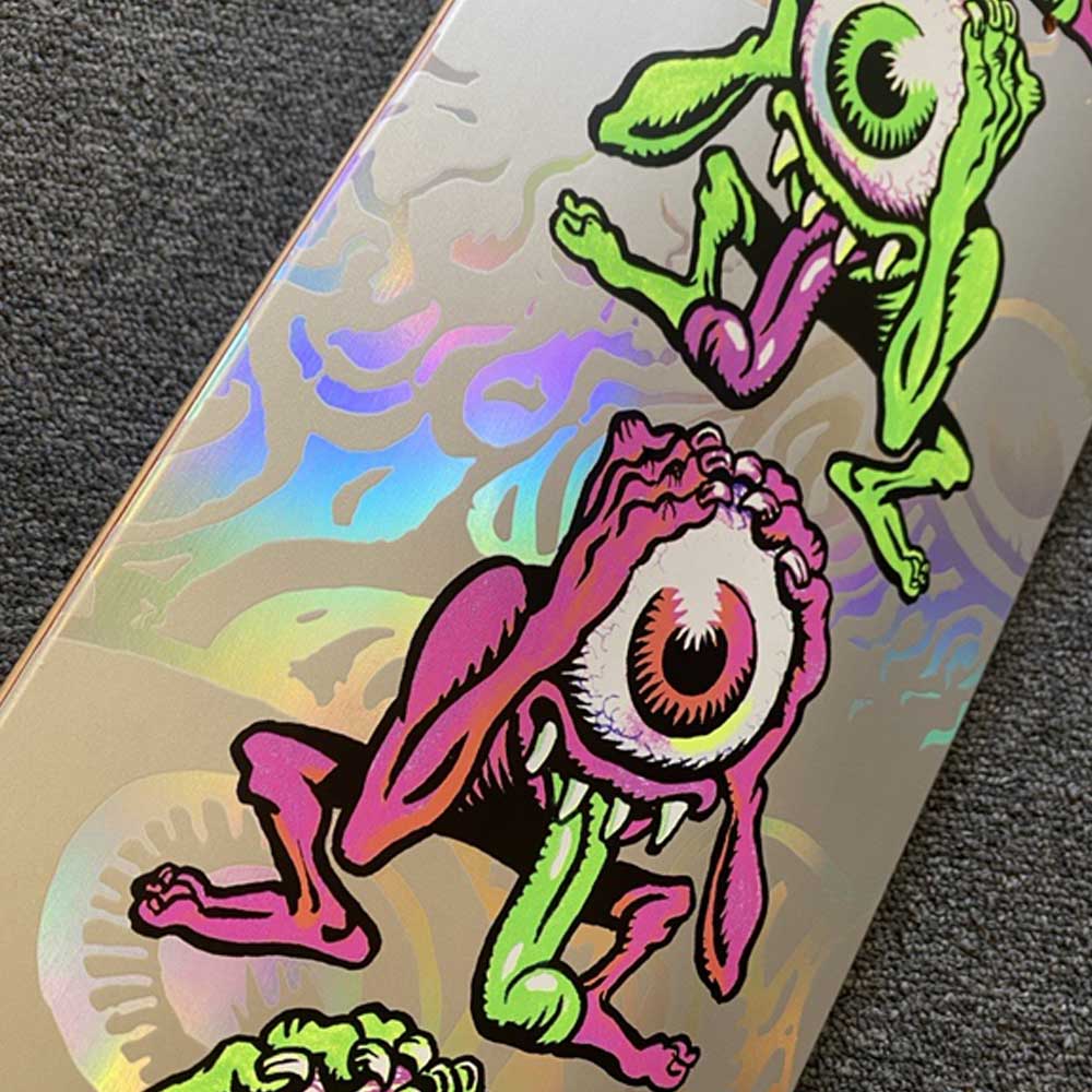 A close-up of the Santa Cruz Eyegore Prismatic Feel 8in x 30.5in skateboard deck, featuring green and pink cartoon monsters with one eye, sharp teeth, and tongues on a shiny holographic background.