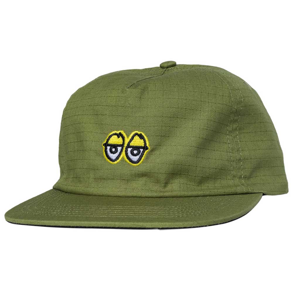 The Eyes Ripstop Olive Krooked Skateboards Adjustable Snapback Hat showcases a small embroidered patch of cartoon eyes, outlined in yellow and black, on the front. Made from durable ripstop olive fabric, this cap features a flat brim and visible stitching details.