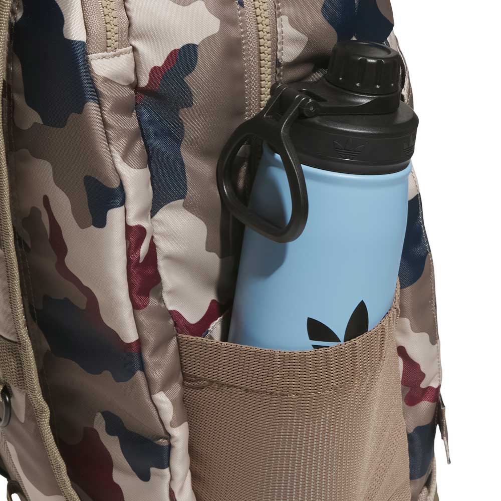 A blue water bottle with a black cap sits in the side pocket of the FSBS Backpack KS6815 Khaki Camo from adidas Skateboarding, showcasing its stylish camouflage pattern and practical design.