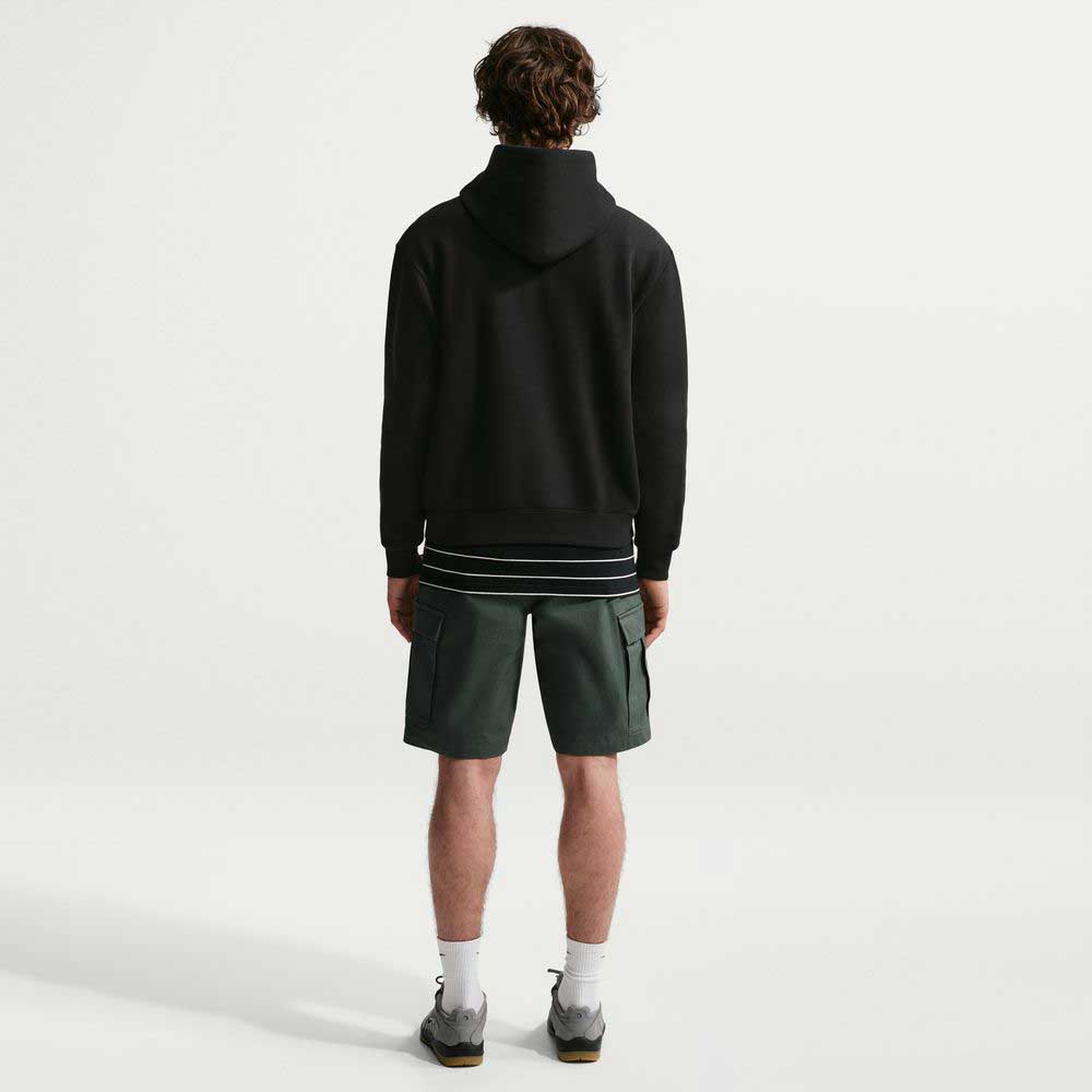 A person faces away from the camera, wearing a Nike SB Fleece Pullover Black White IF0655010 hoodie, dark green shorts, white socks, and sneakers against a plain white background.