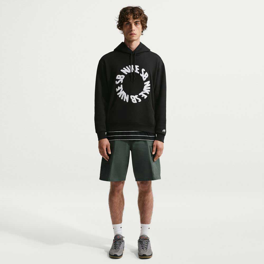 A young man stands facing forward against a plain white background, wearing the Fleece Pullover Black White IF0655010 Hoodie from Nike SB, paired with green shorts, white socks, and gray sneakers.