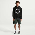 A young man stands facing forward against a plain white background, wearing the Fleece Pullover Black White IF0655010 Hoodie from Nike SB, paired with green shorts, white socks, and gray sneakers.