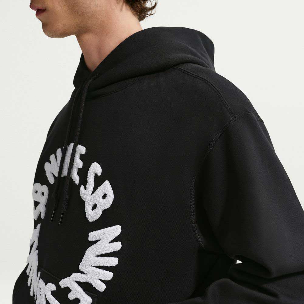 A person wears the Nike SB Fleece Pullover Black White IF0655010 Hoodie, featuring white textured letters in a partial circle on the front. The face is partially out of frame against a plain, light background.
