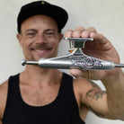A smiling man in a black tank top and cap holds a set of Independent 144 Stage 11 Forged Titanium Chris Joslin Sticker Slap Pro Polished Silver Standard Trucks (set of two) towards the camera, with a blurred background.