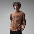 A person with curly hair stands against a plain gray background, looking slightly to the side with hands in pockets, wearing the Fork in the Road Former Tee Washed Bark—crafted from premium jersey cotton with a unique graphic screen print—and black pants.