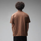 A person with curly hair is standing with their back to the camera, wearing the Fork in the Road Former Tee in Washed Bark—crafted from premium jersey cotton—and black pants against a plain gray background.