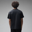 A person with curly hair, dressed in the Engineered Former Tee Washed Black made from 200GSM cotton and dark pants, stands with their back to the camera against a plain gray background.