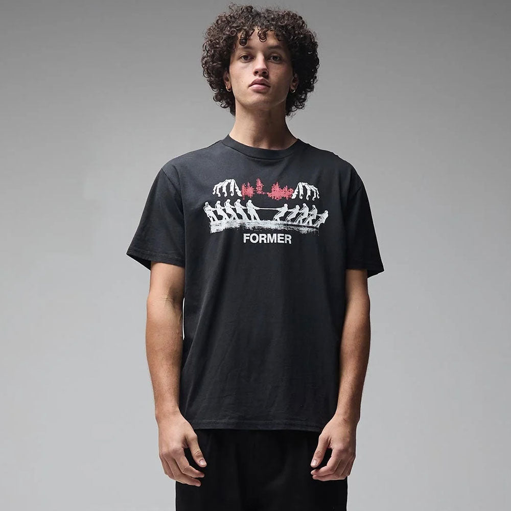 A young man with curly hair stands facing forward, wearing the Engineered Former Tee Washed Black—a vintage-style T-shirt made from 200GSM cotton, featuring a screen-printed graphic design with the word “FORMER” below it—against a plain gray background.