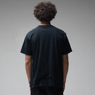 A person with curly hair is standing with their back to the camera, wearing the Mind Glass Former Tee Black—a plain black 200GSM cotton T-shirt—against a neutral gray background.
