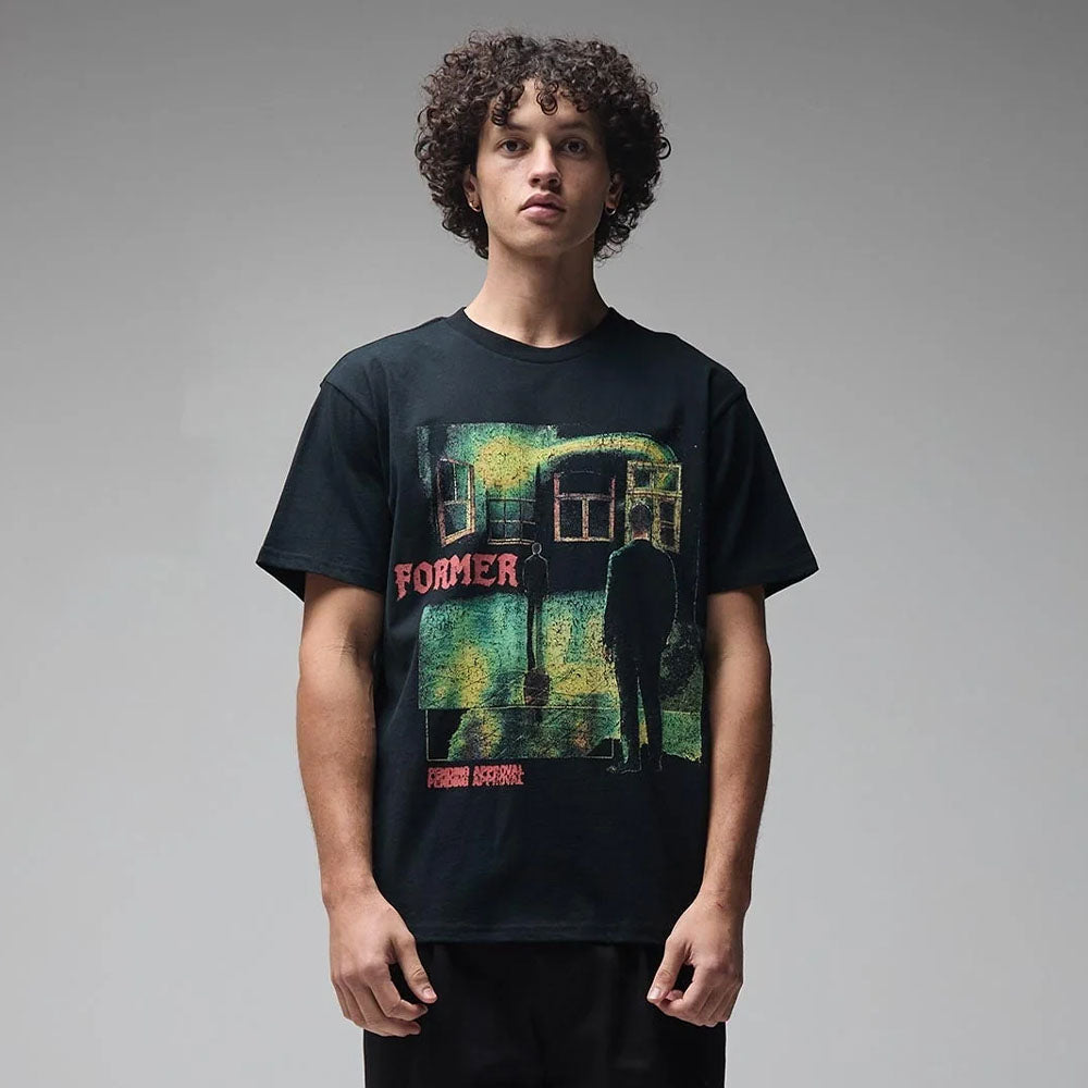 A person with curly hair stands against a plain gray background, wearing the Mind Glass Former Tee Black—a 200GSM cotton T-shirt featuring a colorful screen print of a silhouetted figure and the word “FORMER” in red.