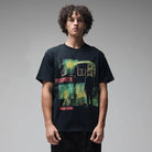 A person with curly hair stands against a plain gray background, wearing the Mind Glass Former Tee Black—a 200GSM cotton T-shirt featuring a colorful screen print of a silhouetted figure and the word “FORMER” in red.