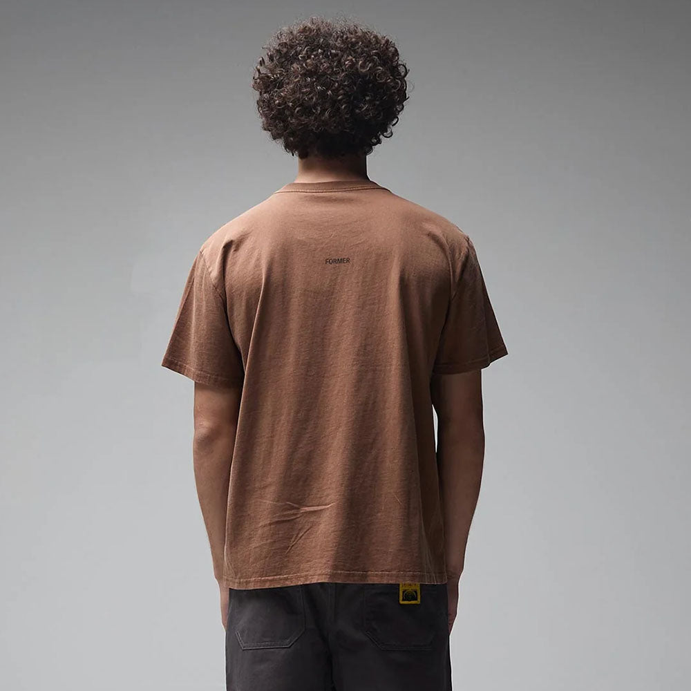 Seen from behind against a plain gray backdrop, a person with curly hair wears the Crux Overdye Former Tee in Washed Bark—crafted from premium jersey cotton—paired with dark pants.