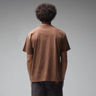 Seen from behind against a plain gray backdrop, a person with curly hair wears the Crux Overdye Former Tee in Washed Bark—crafted from premium jersey cotton—paired with dark pants.