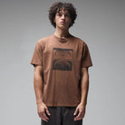 A young man with curly hair wears the Crux Overdye Former Tee in Washed Bark—a brown vintage-style t-shirt crafted from premium jersey cotton, featuring a bold graphic screen print—paired with dark pants, standing against a plain gray background and facing forward with a neutral expression.