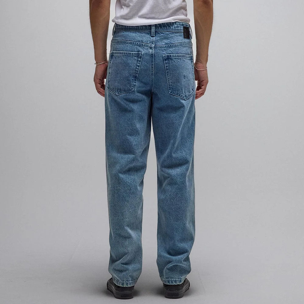 A person wearing the Crux Denim Pant Washed Blue Former—light blue, straight-fit jeans made from 100% cotton denim—and a white t-shirt stands facing away from the camera against a plain gray background.