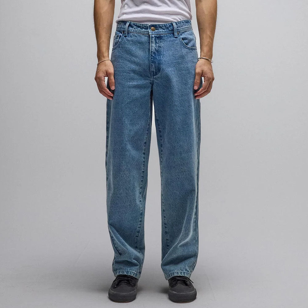 A person is wearing the Crux Denim Pant Washed Blue Former—loose-fitting jeans crafted from 100% cotton denim—paired with a white t-shirt and black shoes, standing against a plain gray background with arms relaxed at their sides.