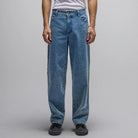 A person is wearing the Crux Denim Pant Washed Blue Former—loose-fitting jeans crafted from 100% cotton denim—paired with a white t-shirt and black shoes, standing against a plain gray background with arms relaxed at their sides.