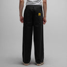 A person stands facing away, wearing loose black Reynolds Work Pant Black Former with two back pockets, one featuring a small yellow patch, paired with a white T-shirt and beige sneakers, against a plain gray background.