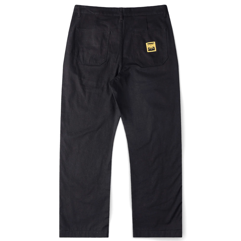 A pair of Reynolds Work Pant Black Former is displayed from the back, showcasing two rear pockets. One pocket is adorned with a small yellow and black rectangular patch label, adding a distinctive touch to these baggy work pants inspired by the Dane Reynolds style.