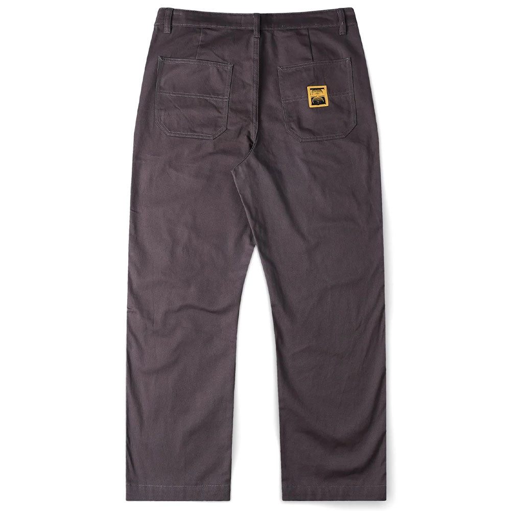 The Reynolds Work Pant Gunmetal Former is shown from the back in dark gray cotton twill, featuring two large rear pockets—one adorned with a small yellow rectangular patch with a black logo. These baggy work pants are inspired by Dane Reynolds’ signature relaxed style.