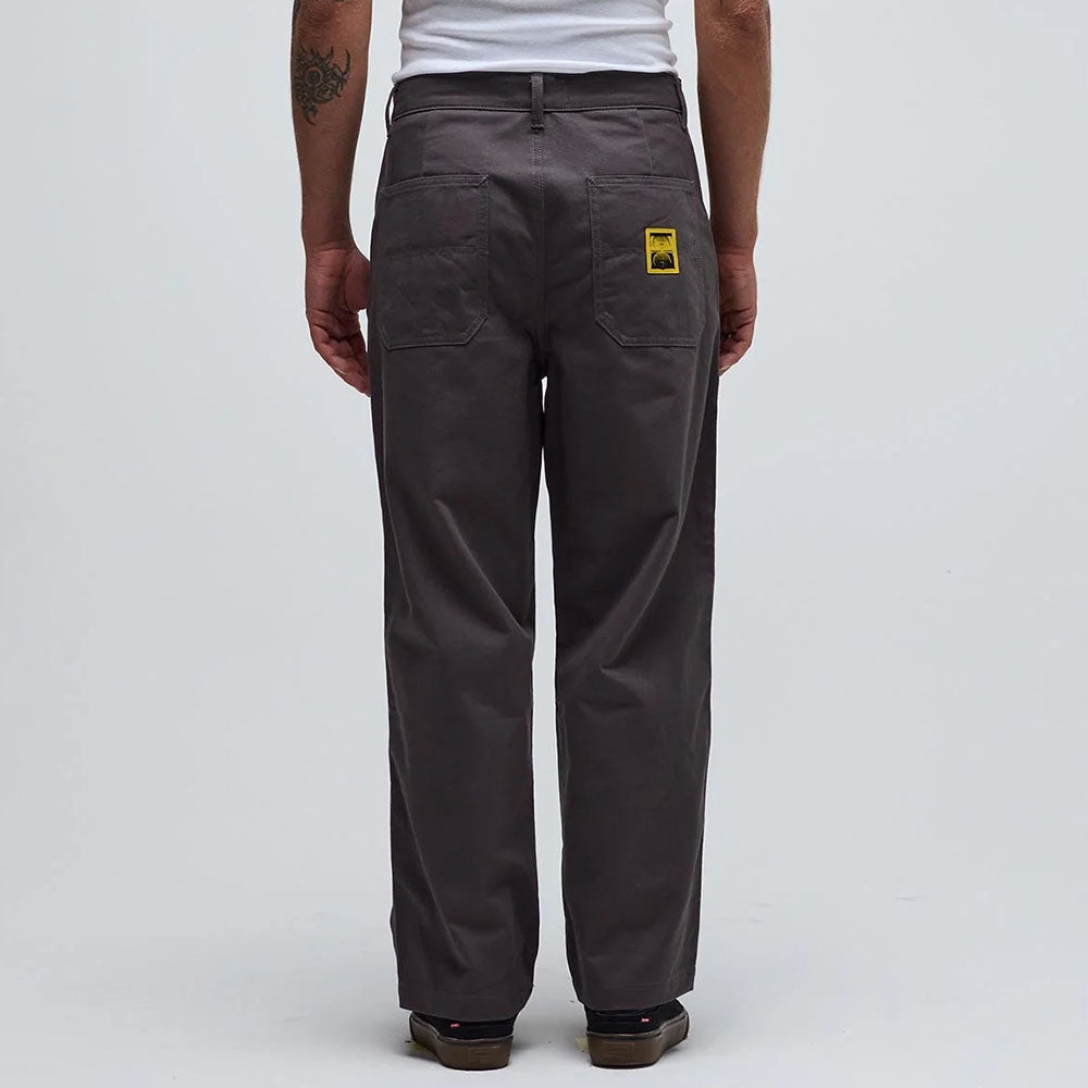 A person stands facing away in the Reynolds Work Pant Gunmetal Former, featuring a gray baggy fit with two back pockets and a yellow patch on the right pocket, paired with a white shirt against a plain light background.