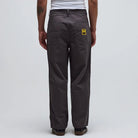 A person stands facing away in the Reynolds Work Pant Gunmetal Former, featuring a gray baggy fit with two back pockets and a yellow patch on the right pocket, paired with a white shirt against a plain light background.