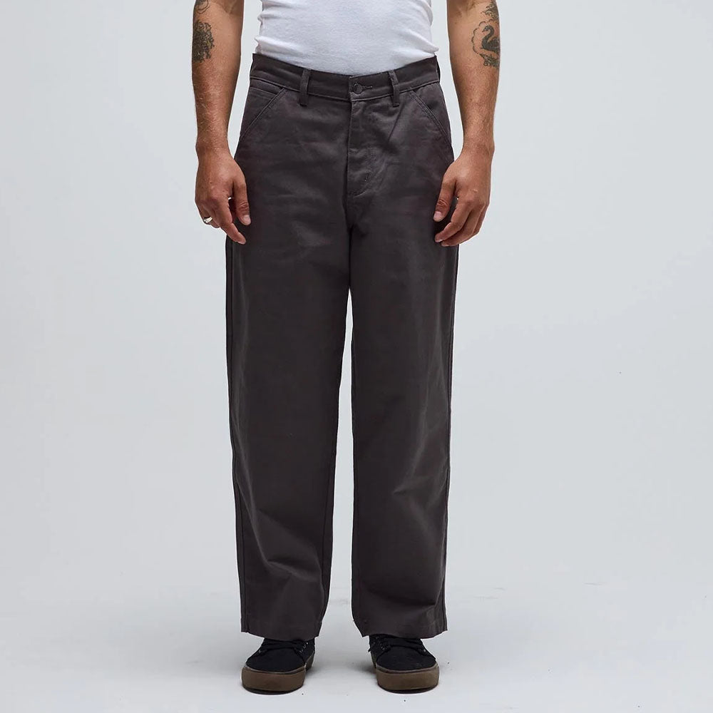 A person stands facing forward, arms relaxed at their sides, wearing the Reynolds Work Pant Gunmetal Former—loose-fitting dark gray cotton twill pants paired with a white T-shirt. The background is plain and light-colored.
