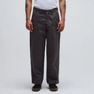 A person stands facing forward, arms relaxed at their sides, wearing the Reynolds Work Pant Gunmetal Former—loose-fitting dark gray cotton twill pants paired with a white T-shirt. The background is plain and light-colored.