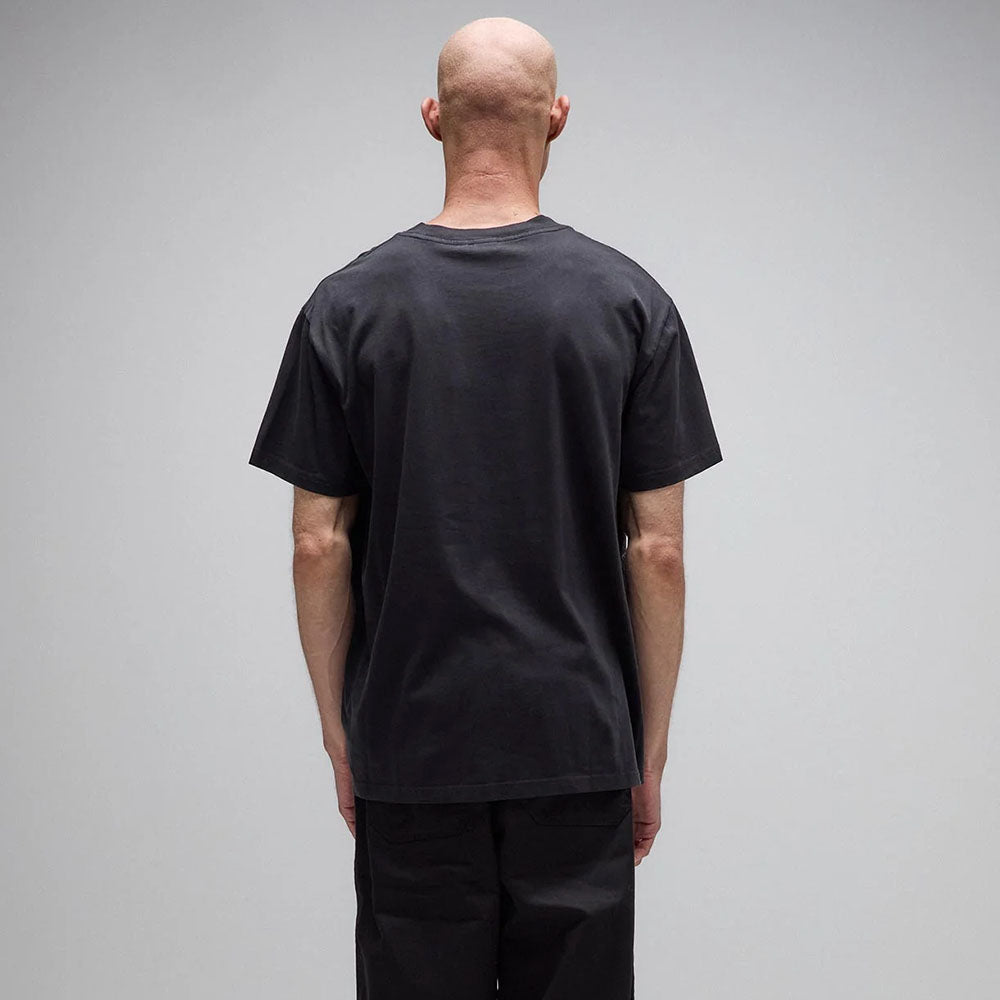 A bald person stands facing away from the camera, wearing the Legacy Overdye Former Tee in washed black—crafted from 200GSM cotton—and black pants, set against a plain light gray background.
