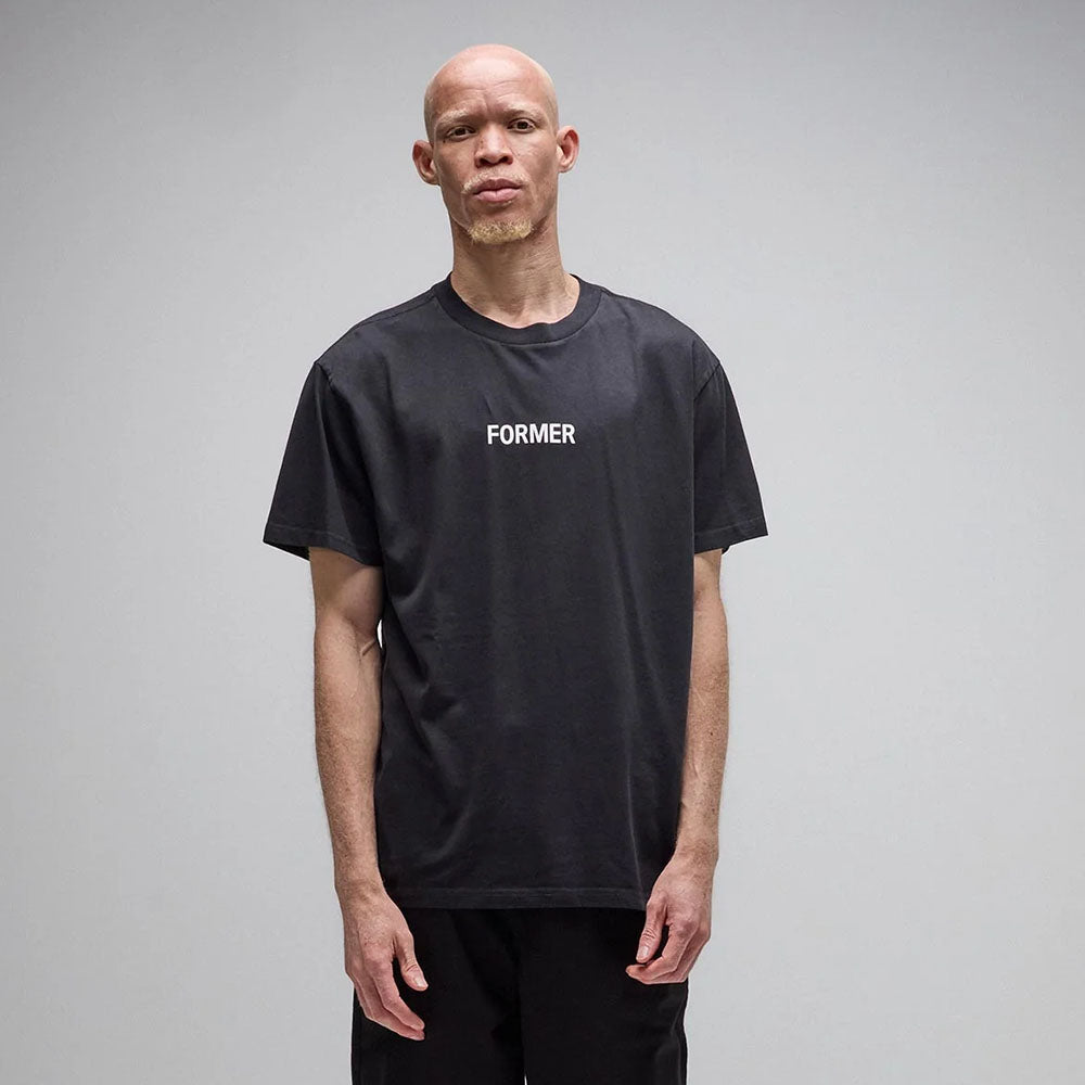 A man with short hair stands against a plain background, wearing the Legacy Overdye Former Tee in washed black. This vintage-style T-shirt is crafted from premium 200GSM cotton and features the word FORMER screen printed in white across the chest.