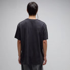 A person stands with their back to the camera, wearing the Crux Overdye Former Tee in Washed Black—crafted from 200GSM cotton for a vintage, loose fit—paired with dark pants against a plain gray background.