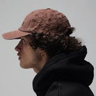 A person with curly hair wearing the Alleviate Cap Former Hat in Bark, a black hoodie layered over a vintage T-shirt, and a small hoop earring, shown in profile against a plain gray background.