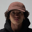 A person with curly hair faces forward against a plain background, wearing the Alleviate Cap Former Hat Bark—a brown cap embroidered with “FORMER”—along with gold hoop earrings and a black hoodie made from soft 200GSM cotton.
