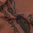 A close-up of the Fork in the Road Former Tee in Washed Bark, showcasing its premium brown jersey cotton fabric with a black screen print of two cars colliding and partial, bold text reading “ENDING APPRO” and “FOR.” The slightly wrinkled material evokes the feel of a vintage T-shirt.