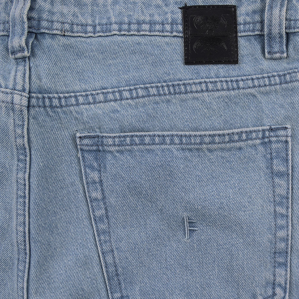 Close-up of the Crux Denim Pant Washed Blue Former, featuring light blue straight fit jeans with a back pocket, belt loop, and a black leather patch on the waistband. Made from 100% cotton denim with a vintage wash, the back pocket is detailed with a small embroidered F near the top.