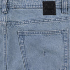 Close-up of the Crux Denim Pant Washed Blue Former, featuring light blue straight fit jeans with a back pocket, belt loop, and a black leather patch on the waistband. Made from 100% cotton denim with a vintage wash, the back pocket is detailed with a small embroidered F near the top.