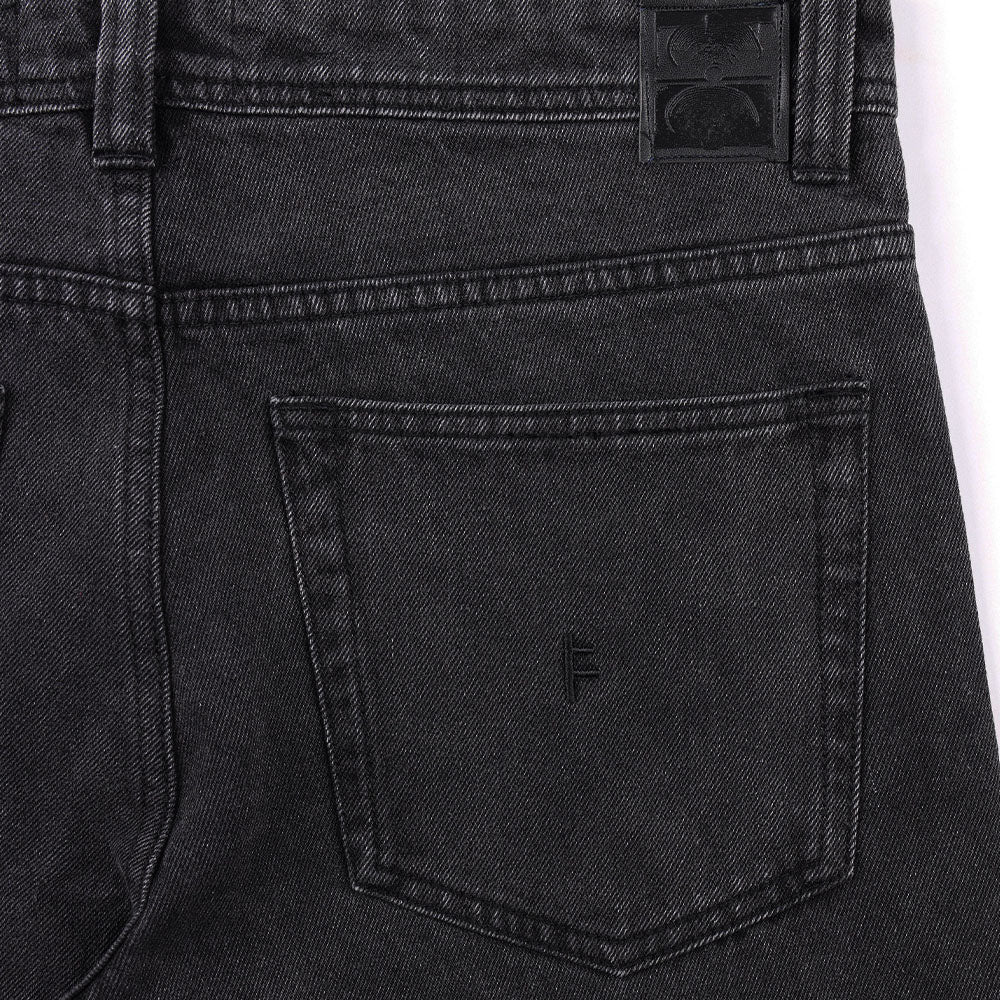 Close-up of the back pocket on a pair of Crux Denim Pant Washed Black Former jeans, showcasing visible stitching details and a subtle embroidered logo near the center of the pocket. Made from 100% cotton denim in a straight fit, these pants are finished with a black leather patch above the pocket.