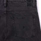 Close-up of the back pocket on a pair of Crux Denim Pant Washed Black Former jeans, showcasing visible stitching details and a subtle embroidered logo near the center of the pocket. Made from 100% cotton denim in a straight fit, these pants are finished with a black leather patch above the pocket.