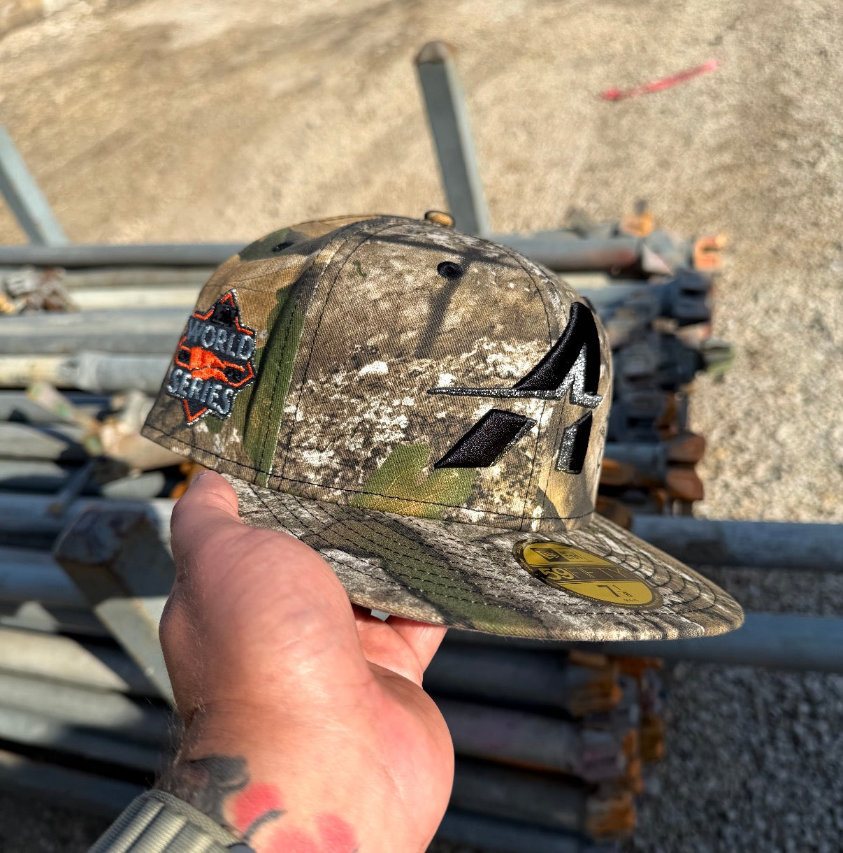 A hand holds The Blackland Apex WS22 Houston Astros New Era Hat 5950 Fitted, featuring a camouflage design, a World Series patch, and a gold sticker on the brim. Metal pipes and gravel are in the background.