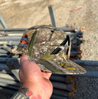 A hand holds The Blackland Apex WS22 Houston Astros New Era Hat 5950 Fitted, featuring a camouflage design, a World Series patch, and a gold sticker on the brim. Metal pipes and gravel are in the background.