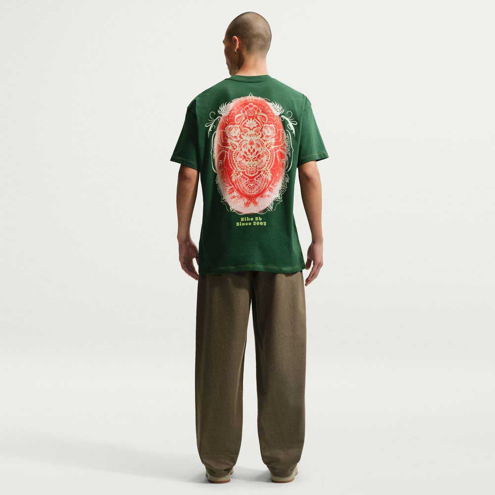 A person with a shaved head wears the Nike SB Gorge Green Som Tum Dunk Tee IH1478341, a roomy midweight cotton T-shirt with a large red and white circular back graphic, paired with loose brown pants against a white background.