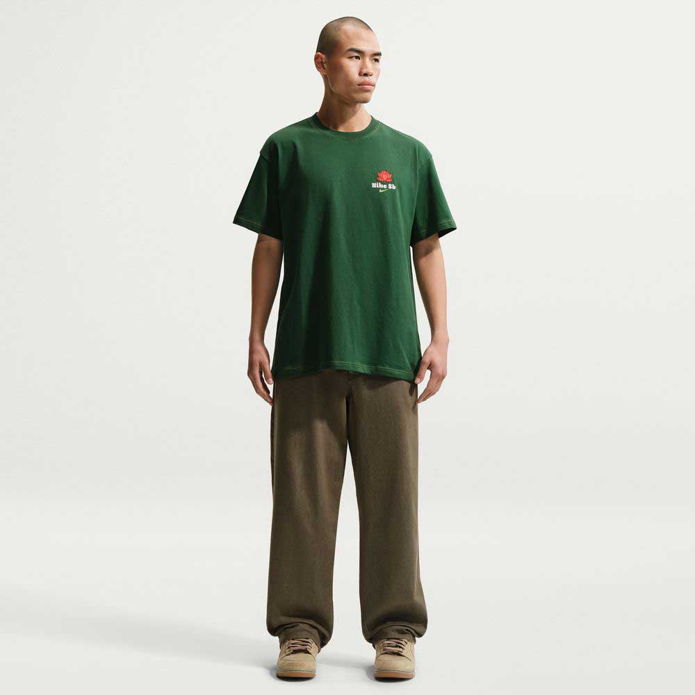 A person with a shaved head faces forward, wearing the Nike SB Gorge Green Som Tum Dunk Tee IH1478341—a roomy fit, midweight skateboarding T-shirt—paired with baggy olive-green pants and beige sneakers against a white background.