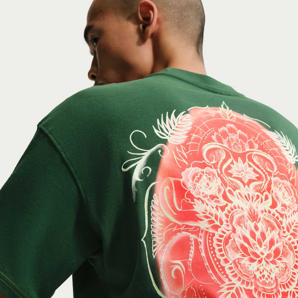 Someone wears the Gorge Green Som Tum Dunk Tee (IH1478341), a Nike SB Skateboarding T-shirt in dark green cotton, featuring a bold pink and white floral back design, set against a plain light background.