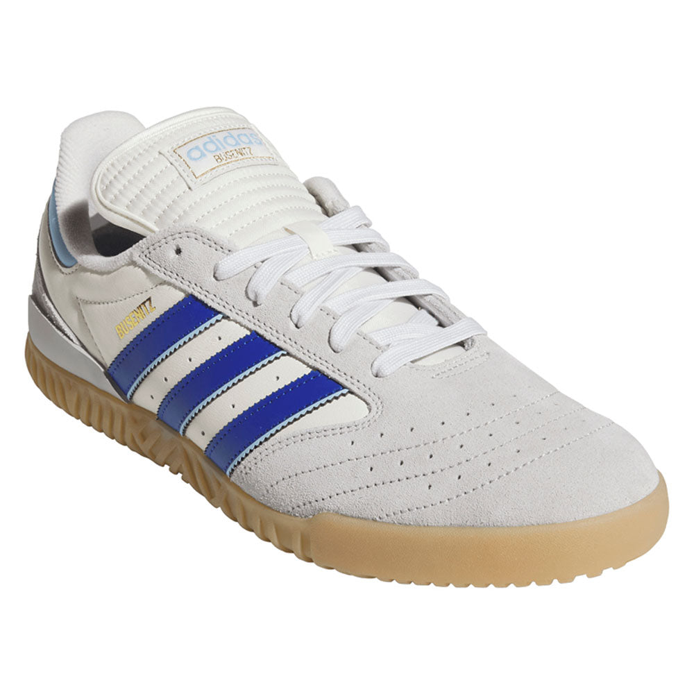 A gray adidas Busenitz Indoor Super skateboarding shoe, featuring royal blue stripes, gold accents, and a gum sole. The suede footwear is shown at an angle highlighting the side, top, and front, blending classic style with performance elements drawn from the iconic silhouette.
