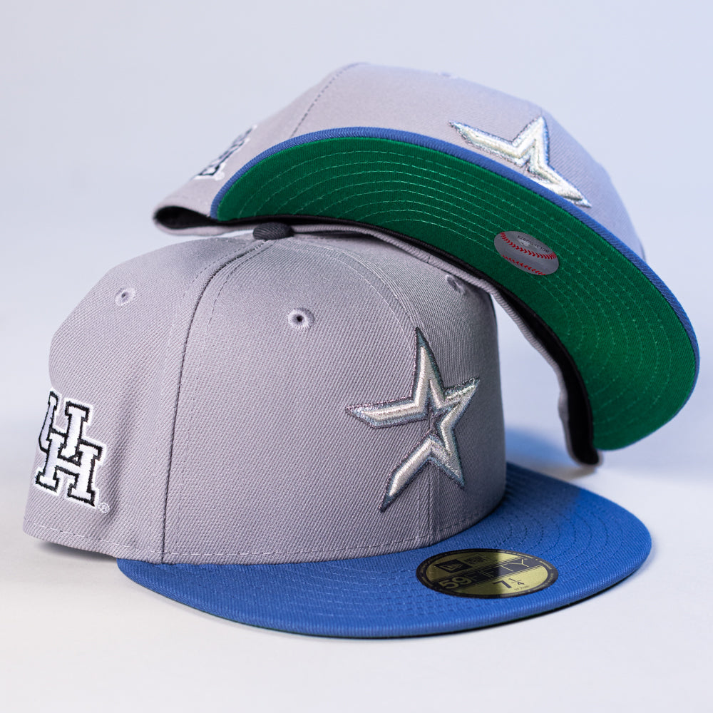 Two light gray and blue Peoples Champ Gray Blue Houston Cougars New Era Hat 5950 Fitted caps by New Era, each featuring silver star and H logo patches—one cap resting on top of the other; the upper cap displays its classic green underbrim and sticker, showcasing iconic Houston Cougars style.