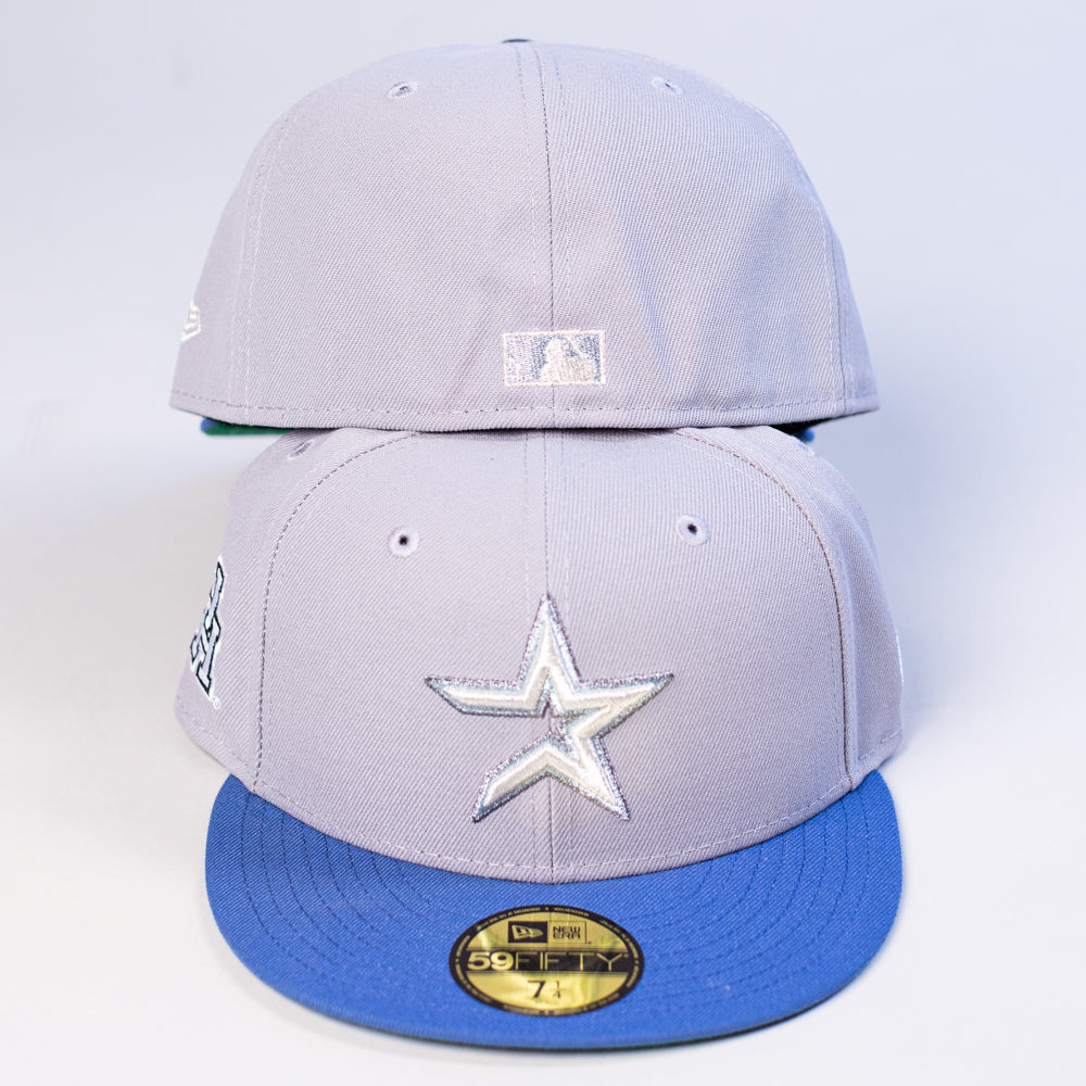 Two light gray Peoples Champ Gray Blue Houston Cougars New Era Hat 5950 Fitted caps are stacked, each featuring blue flat brims and visible stitching. The front cap displays a metallic star logo, while the back cap shows a small white emblem on the rear—making these hats perfect for Houston Cougars fans seeking standout style.